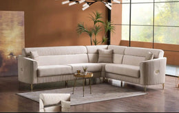 A MODREN L-SHAPED BEIGE SOFA FEATURING VERTICAL TUFTING , SOFT UPHOLSTERY , ELEGANT GOLD  FINISHED METAL LEGS , AND DECORATIVE GOLD FINISHED METAL LEGS AND DECORATIVE GOLD ACCENTS ON THE ARMRESTS. MEAUREMENT  295X235CM