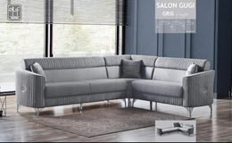 A MODREN L-SHAPED GREY SOFA FEATURING VERTICAL TUFTING , SOFT UPHOLSTERY , ELEGANT SILVER FINISHED METAL LEGS , AND DECORATIVE SILVER FINISHED METAL LEGS AND DECORATIVE SILVER ACCENTS ON THE ARMRESTS. MEAUREMENT  295X235CM